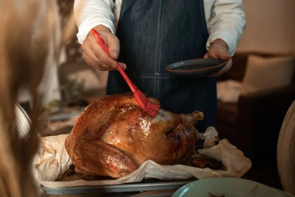 A person wearing an apron glazes a roasted turkey, preparing for a Thanksgiving feast indoors.